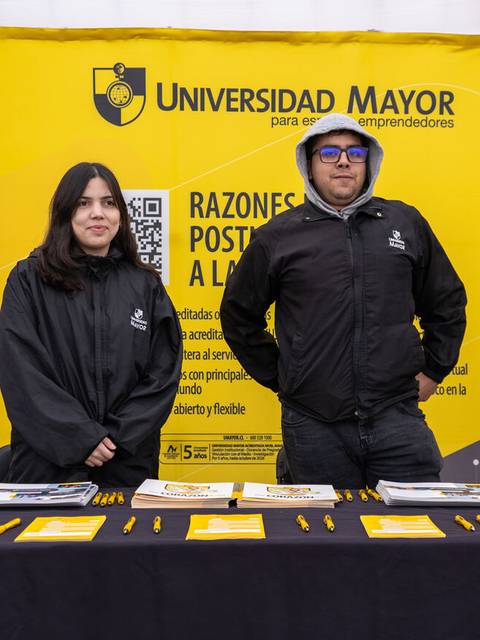 Un par de personas están de pie frente a un stand de la Universidad Mayor con folletos y un cartel informativo.