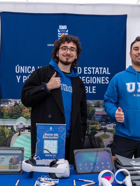 Dos jóvenes posan sonrientes junto a un stand educativo en un evento.