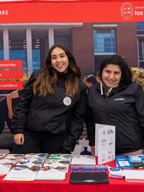 Dos mujeres sonrientes en un stand de la Universidad de los Andes con material informativo sobre programas académicos.