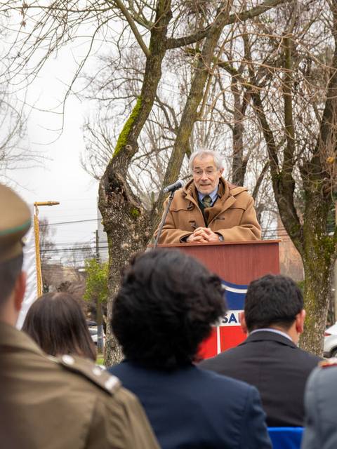 Un hombre mayor habla desde un podio frente a un grupo de personas en un evento al aire libre.