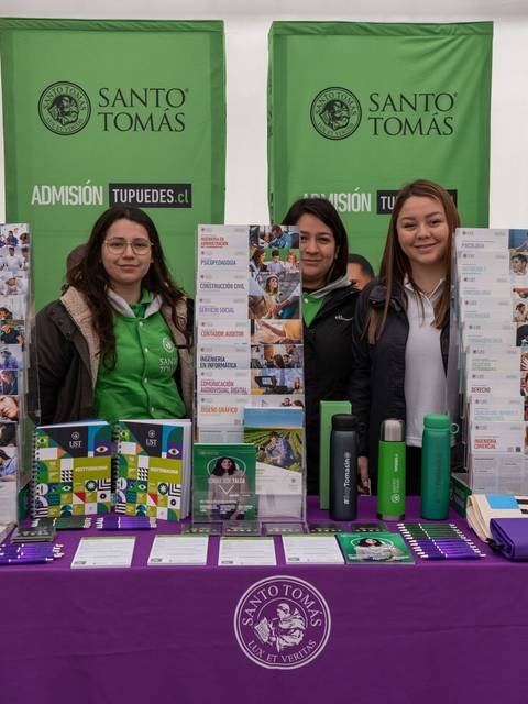 Una feria de admisión universitaria con tres personas posando frente a un stand de Santo Tomás.