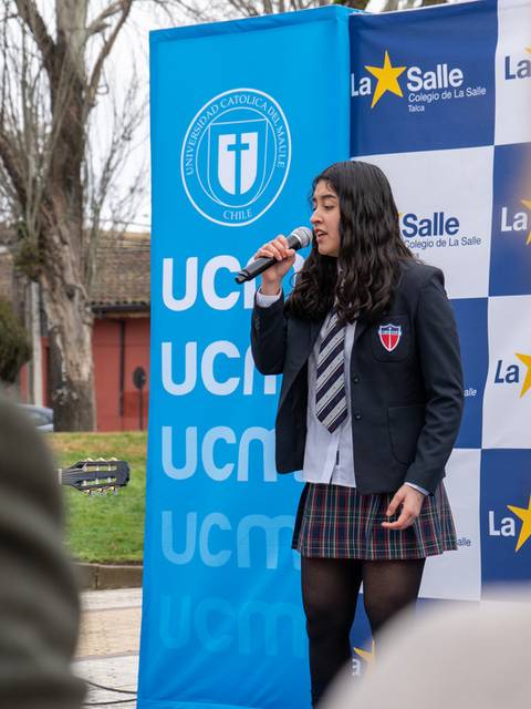 Una estudiante canta en un evento escolar frente a un panel de marcas institucionales.