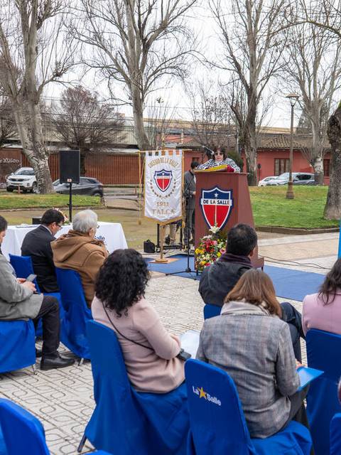 Un grupo de personas sentadas en sillas azules, prestando atención a un evento que se lleva a cabo al aire libre.