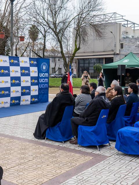 Un evento al aire libre con un orador frente a un público sentado, rodeado de decoraciones y carteles institucionales.