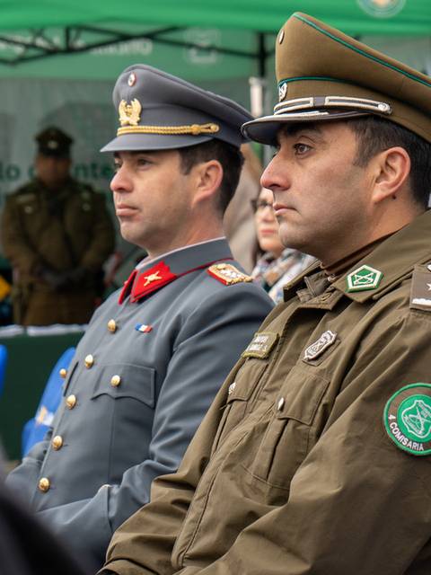 Dos oficiales de uniforme militar asisten a un evento con un público de fondo.