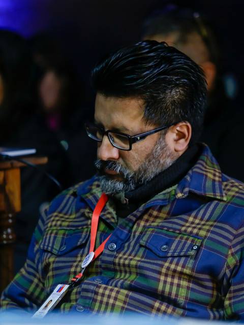 Un hombre con barba y gafas está sentado y parece concentrado en algo, mientras otros asistentes están al fondo.