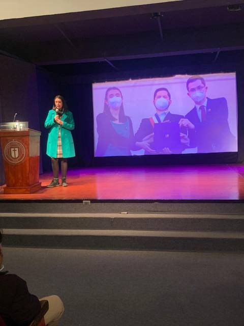 Una mujer habla en un escenario con una presentación proyectada detrás de ella.