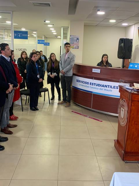 Una ceremonia en un salón donde un grupo de personas está asistiendo a un discurso frente a un escritorio con un panel informativo.