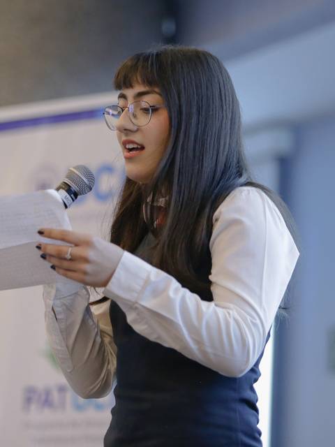 Una joven está hablando frente a un público mientras sostiene un micrófono y un papel en la mano.
