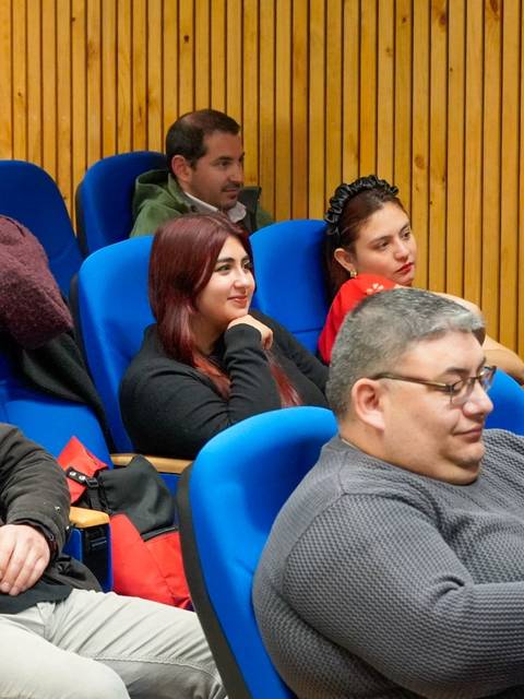 Un grupo de personas sentadas en una sala con asientos azules, prestando atención a un evento.