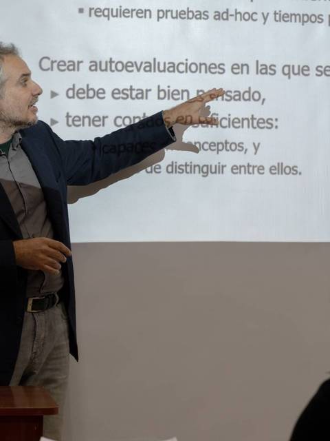 Un hombre está dando una presentación señalando con su mano a una diapositiva en una sala de clase.