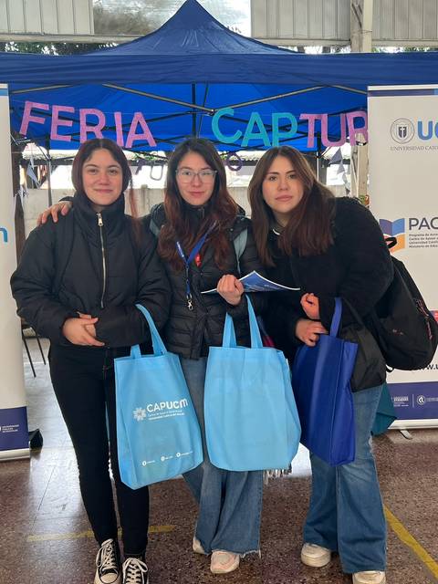 Tres jóvenes posan sonrientes sosteniendo bolsas en una feria educativa.