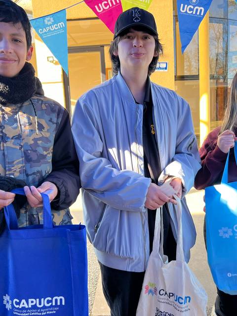 Tres jóvenes sonríen sosteniendo bolsas de tela en un evento al aire libre.