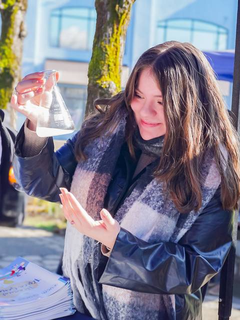 Una joven sostiene un frasco de laboratorio mientras sonríe en un evento al aire libre.