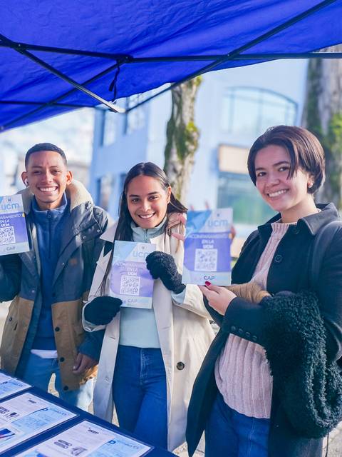 Tres personas sonrientes sosteniendo folletos debajo de un toldo azul.