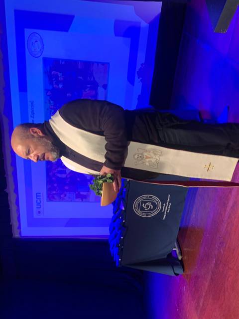 Un hombre vestido con toga sostiene una planta mientras se encuentra de pie sobre un escenario iluminado.