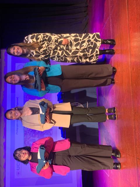 Cuatro mujeres posan en un escenario con trofeos y diplomas sobre un fondo iluminado.