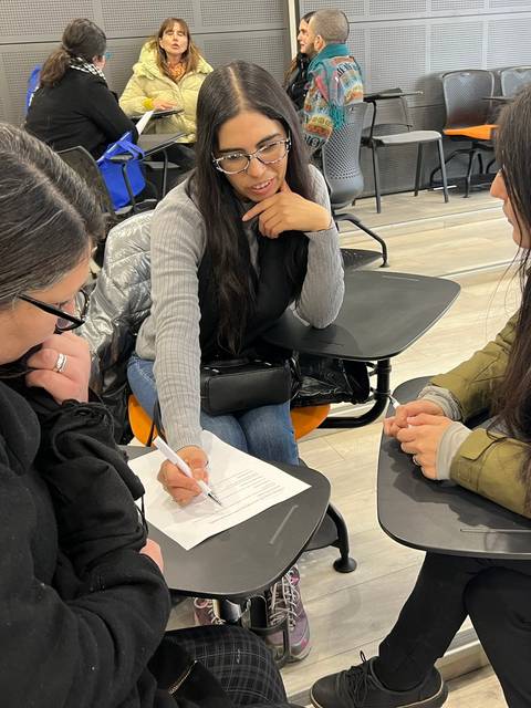 Un grupo de tres jóvenes se sienta en un aula mientras discuten y toman apuntes.