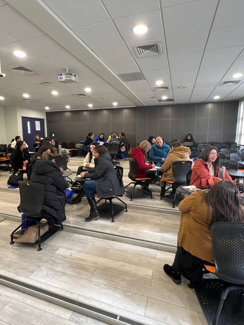 La imagen muestra un aula con varias personas sentadas en grupos, participando en actividades de colaboración.