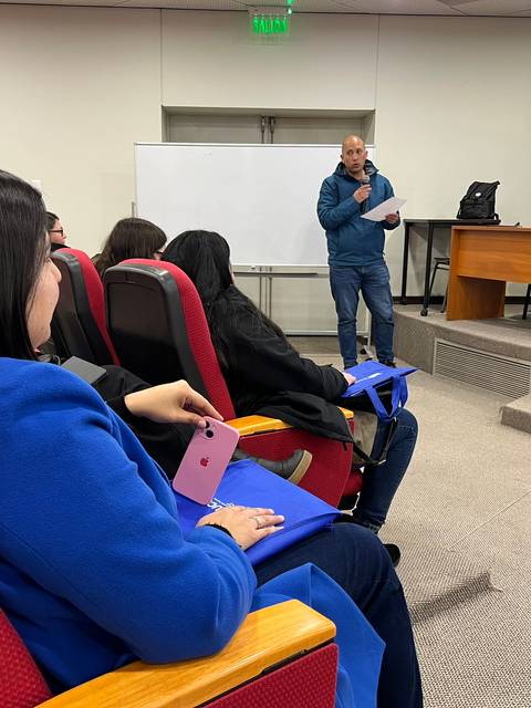 Una reunión en un salón con varias personas sentadas y un orador frente a una mesa.