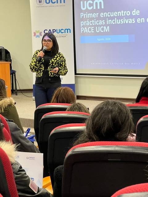 Una mujer habla frente a un grupo de personas en una sala con asientos rojos, durante un encuentro sobre prácticas inclusivas en el aula.