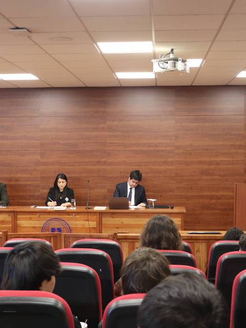 Una conferencia en un aula con público, donde tres personas están sentadas frente a un panel mientras una cuarta persona presenta.
