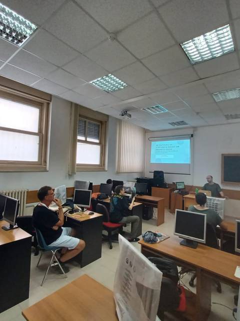 Un aula con varias personas sentadas frente a computadoras mientras un profesor da una lección en la pizarra.