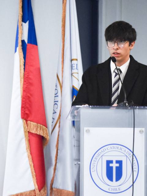 Un joven con gafas se encuentra hablando en un podio frente a dos banderas en un evento formal.