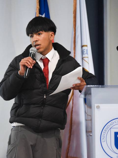 Un joven está hablando en un micrófono durante un evento en una sala con banderas al fondo.