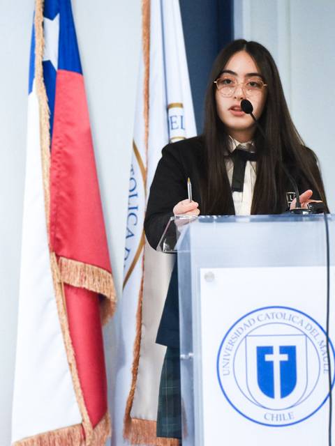 Una mujer joven habla en un podio frente a las banderas de Chile y de la universidad.