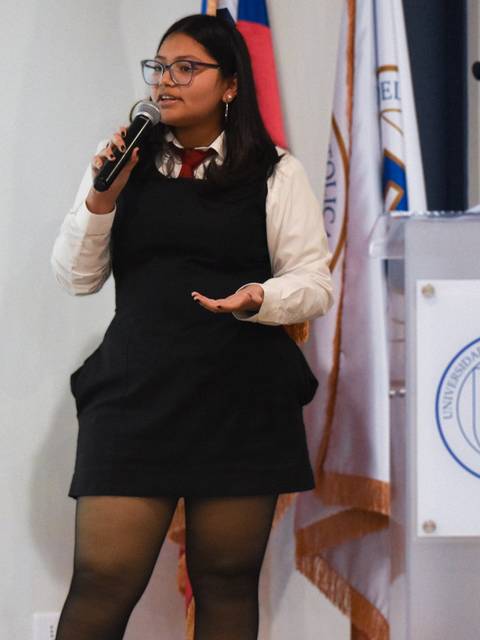 Una joven se encuentra de pie hablando en un micrófono frente a una pantalla en un evento oficial.