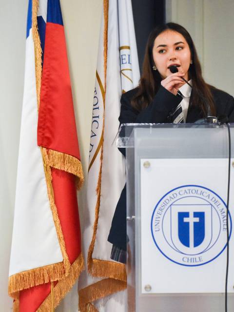 Una joven está hablando en un micrófono frente a un podio decorado con el escudo de una universidad.