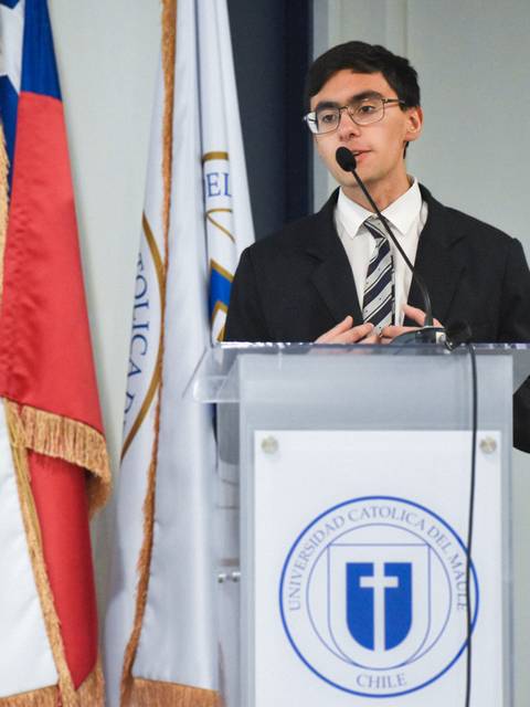 Un joven en traje presenta un discurso frente a un podio con insignias de la universidad y banderas detrás.