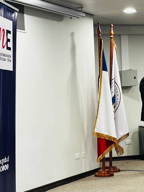 Un hombre está hablando en un evento formal frente a una audiencia, acompañado de algunas banderas y pancartas institucionales.