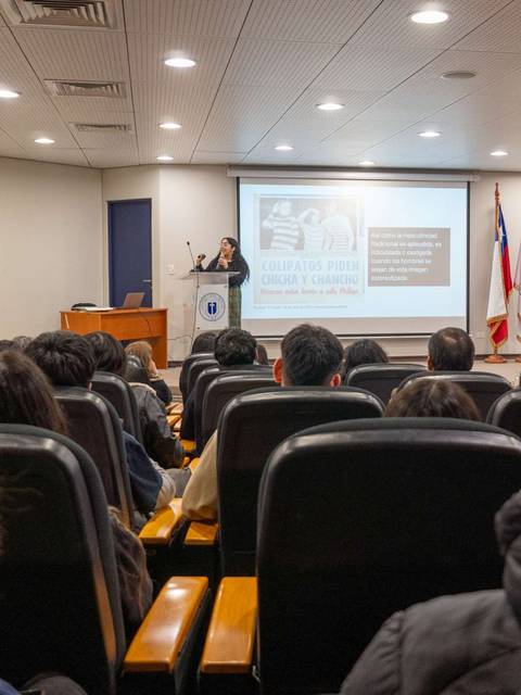 Una conferencia con un presentador frente a una audiencia sentada en un salón.