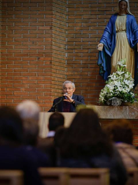 Un hombre habla ante un grupo de personas en un lugar religioso, con una imagen de la Virgen María al fondo.