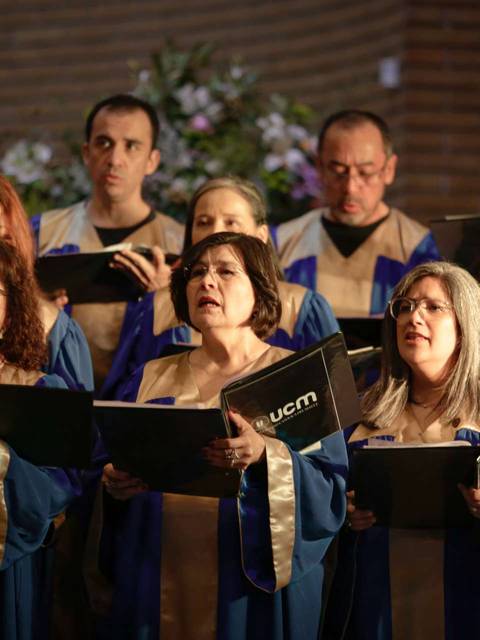 Un grupo de cantantes de un coro se presenta en un escenario, vestidos con túnicas azules y doradas mientras cantan atentamente.