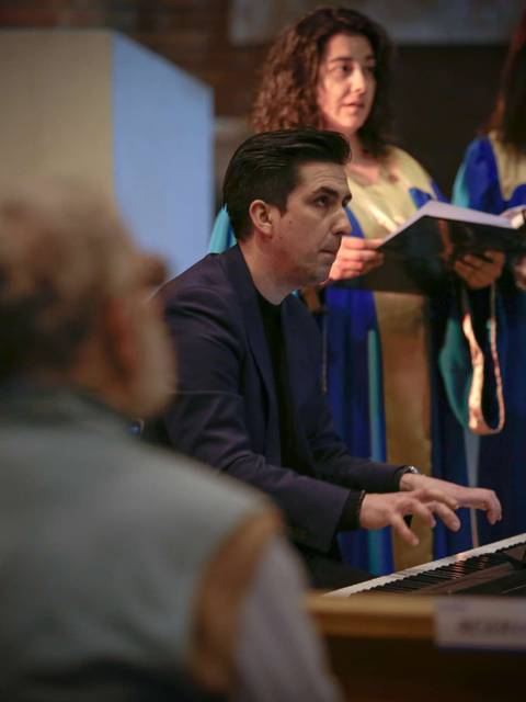 Un músico toca el piano mientras un coro canta en un fondo iluminado.