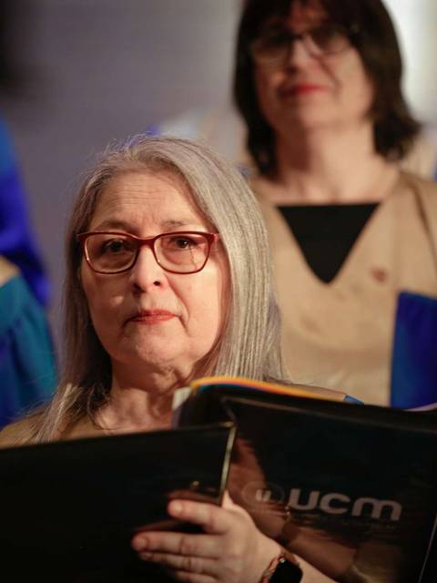 Una mujer con cabello largo y canoso sostiene un libro de partituras mientras observa a un coro en el fondo.