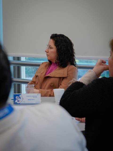 Una mujer con abrigo beige y cabello rizado está escuchando atentamente en una reunión.