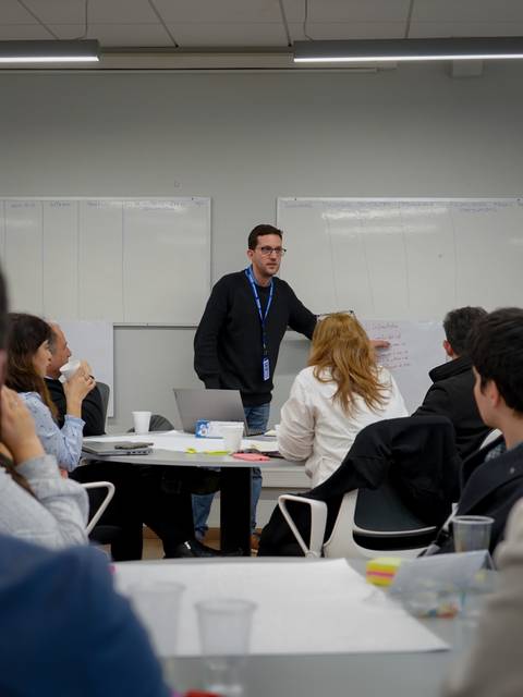 Un grupo de personas participa en una sesión de trabajo en equipo donde un hombre presenta en una pizarra.