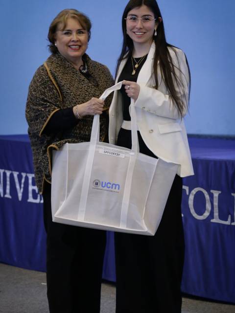 Dos mujeres posan juntas, una de ellas sostiene una bolsa con el logotipo de una universidad.