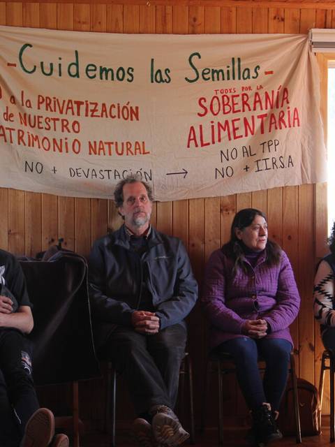 Un grupo de personas se sienta en un salón con un cartel que aboga por la soberanía alimentaria y la protección de las semillas.