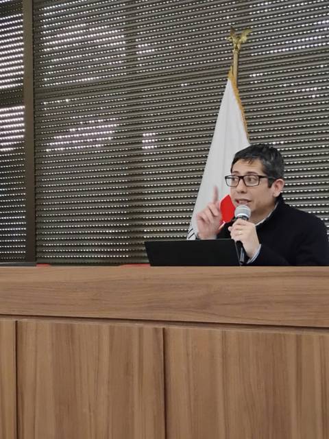 Un hombre está dando una presentación en un Podio con banderas de Chile y Japón al fondo.