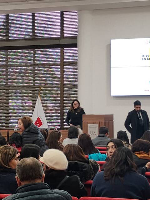 Un grupo de personas se encuentra en un evento educativo en un salón con banderas visibles y una presentación en pantalla.