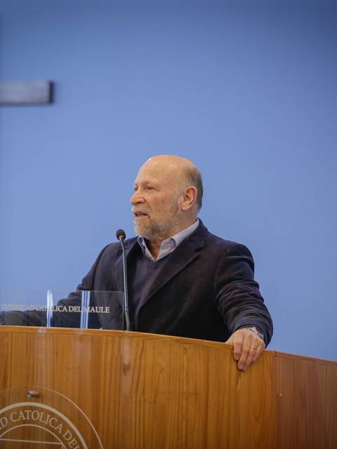 Un hombre con barba habla desde un podio bajo una cruz en un entorno de universidad.