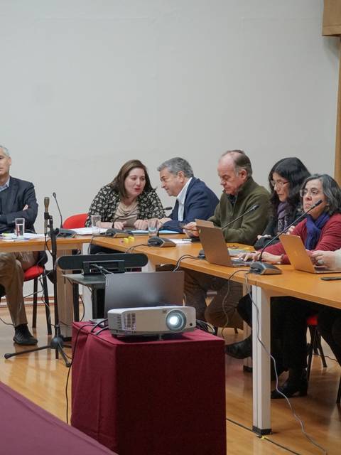 La imagen muestra una reunión con varios participantes sentados alrededor de una mesa en un ambiente formal.
