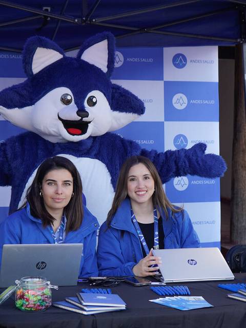 Dos mujeres están sentadas en un stand con computadoras, mientras un disfraz de un animal azul está de pie detrás de ellas con los brazos extendidos.