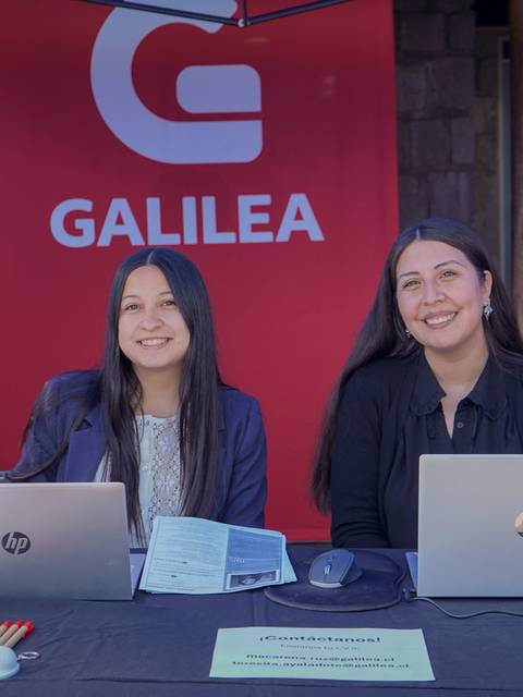Dos mujeres sonrientes están sentadas frente a computadoras en un stand con un fondo rojo que dice 'GALILEA'.