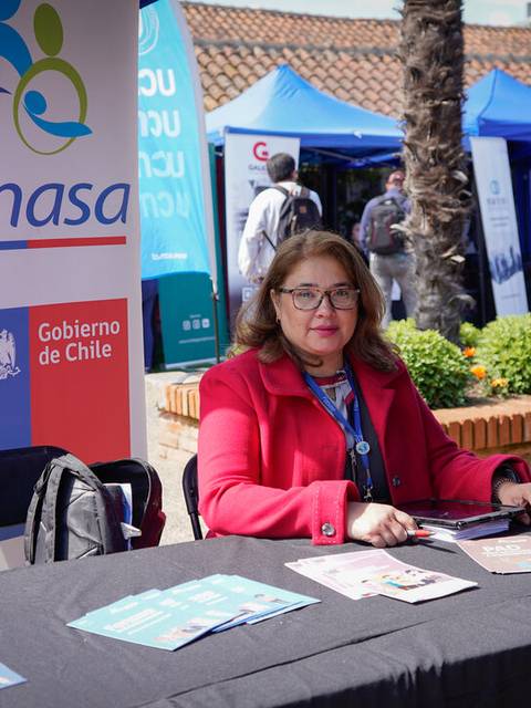 La imagen muestra a una mujer sentada en una mesa con material informativo y un cartel de Fonasa en un evento al aire libre.
